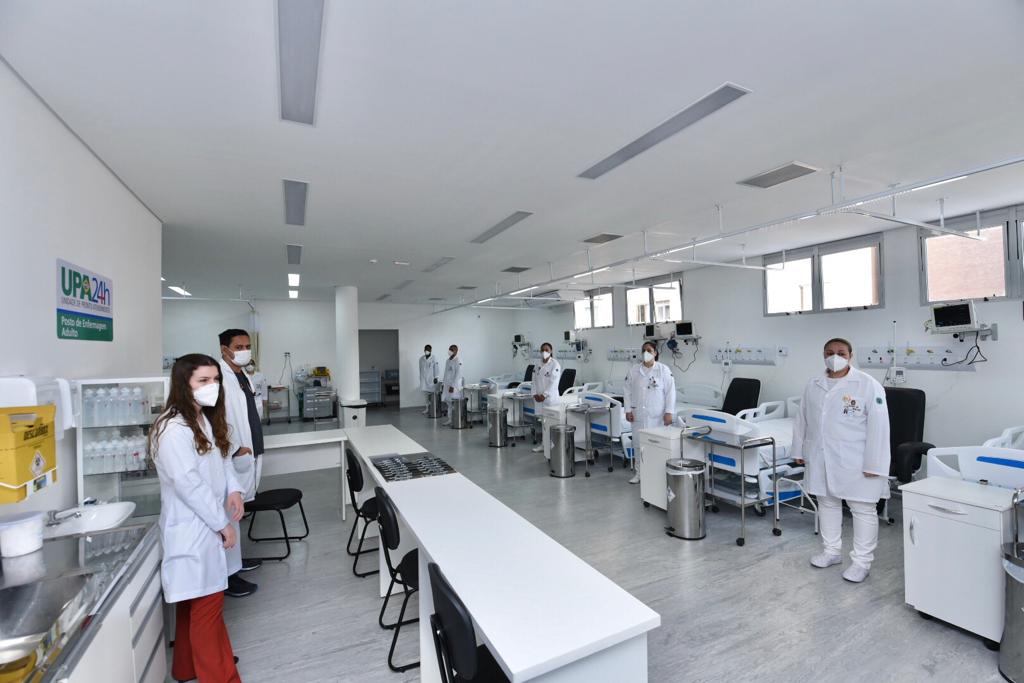 Foto de clínica - A grande sala branca conta com quatro camas de UTI e equipamentos médicos na parede direita, à esquerda temos um ponto de acolhimento dos pacientes, profissionais uniformizados e de máscara ocupam o espaço